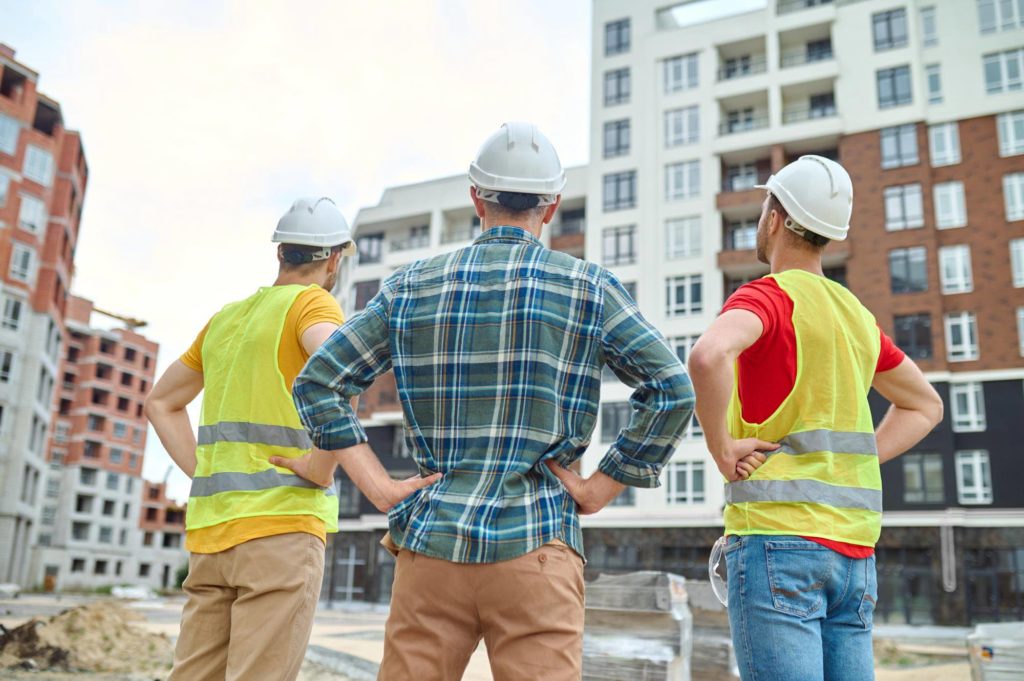 Honoring Our Heroes in Hard Hats 13 - Lunacon Construction Group result back view three men protective helmets holding hands his waist standing construction site looking buildings construction during day